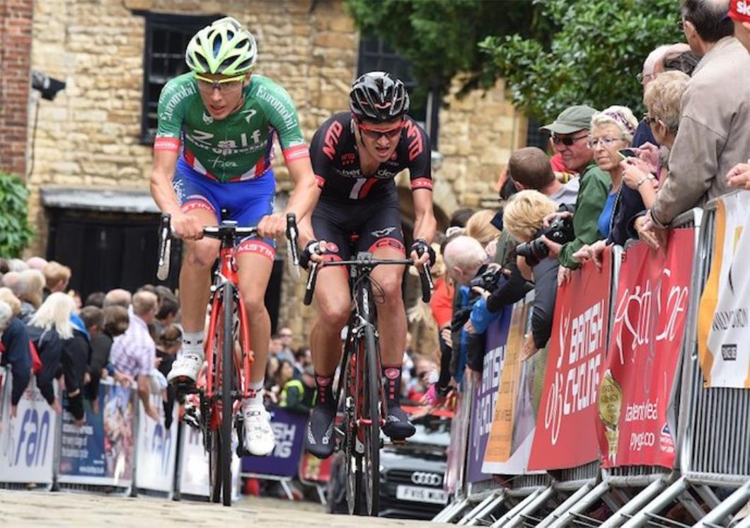 Racing cyclists, two, lead rider in green jersey and blue shorts, following rider in black and red kit, cobbled road, barriers, spectators, traditional house in background, Dan Pearson, pic: Larry Hickmott